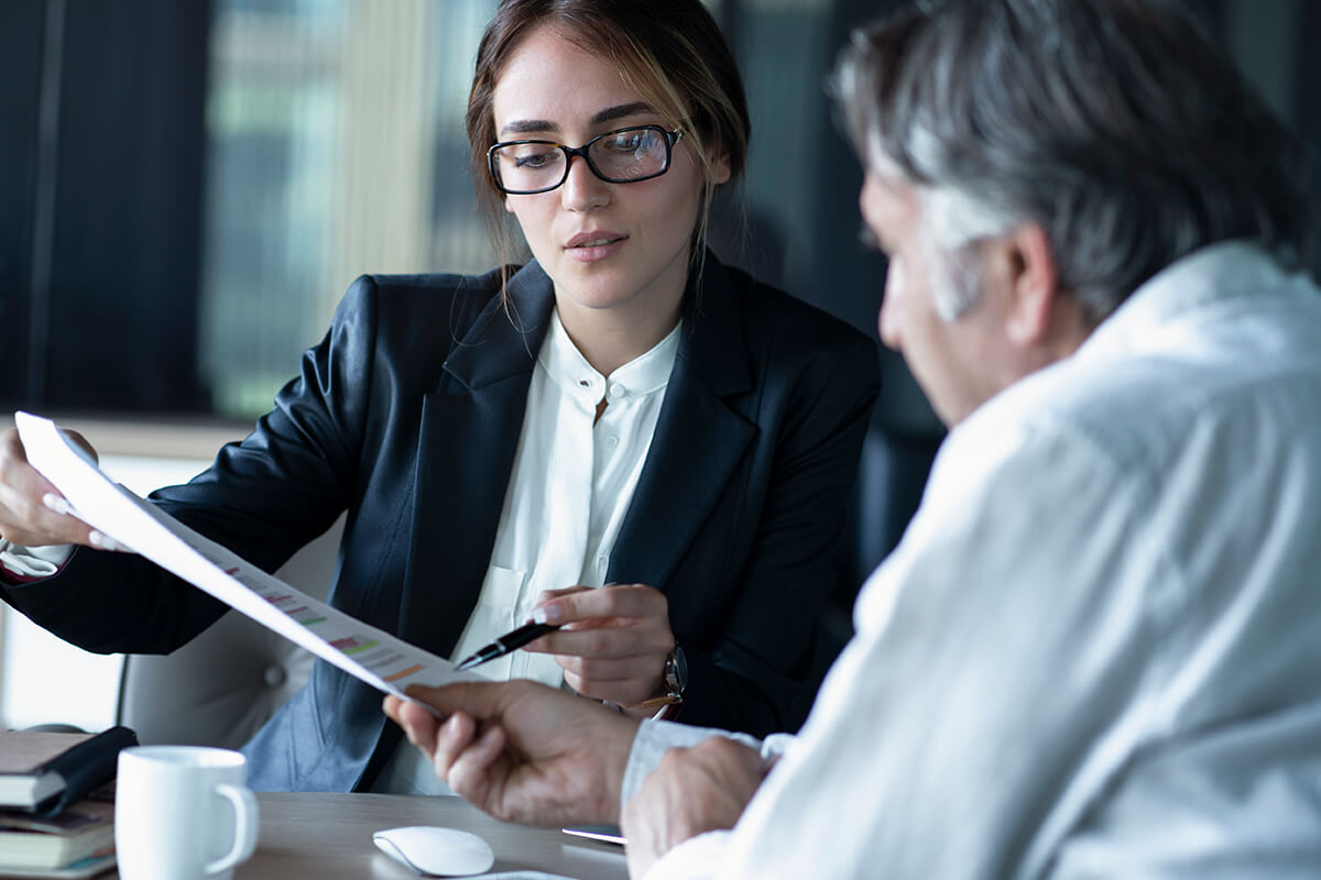 A woman wearing glasses and a dark blazer shows a document to an older man in a white shirt during a discussion in an office setting, likely involving franchise consulting firms.